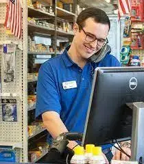 A Man in an auto store, standing behind a desk, on the phone, smiling, looking at a computer screen.