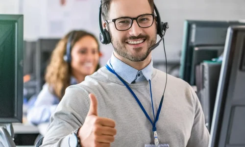 A customer service representative surrounded by data screens, with a smile and a thumbs up and coworker in background.