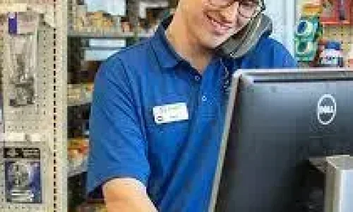 A Man in an auto store, standing behind a desk, on the phone, smiling, looking at a computer screen.